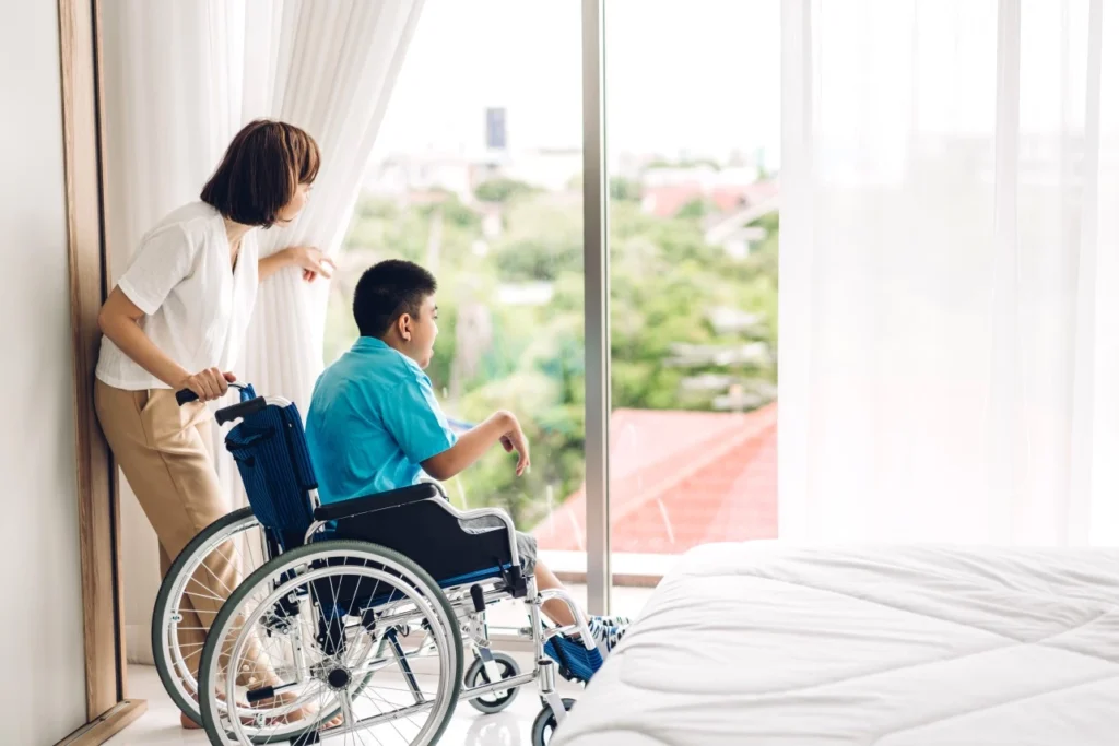 Disabled man in wheelchair with nurse at home in bedroom looking out the window